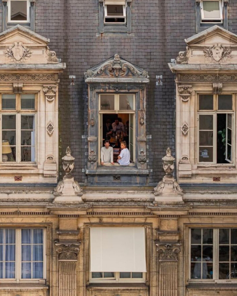Historic Parisian apartment facade with ornate stone window surrounds, slate roof tiles, and people gathered at an open window, capturing a lived-in city moment.