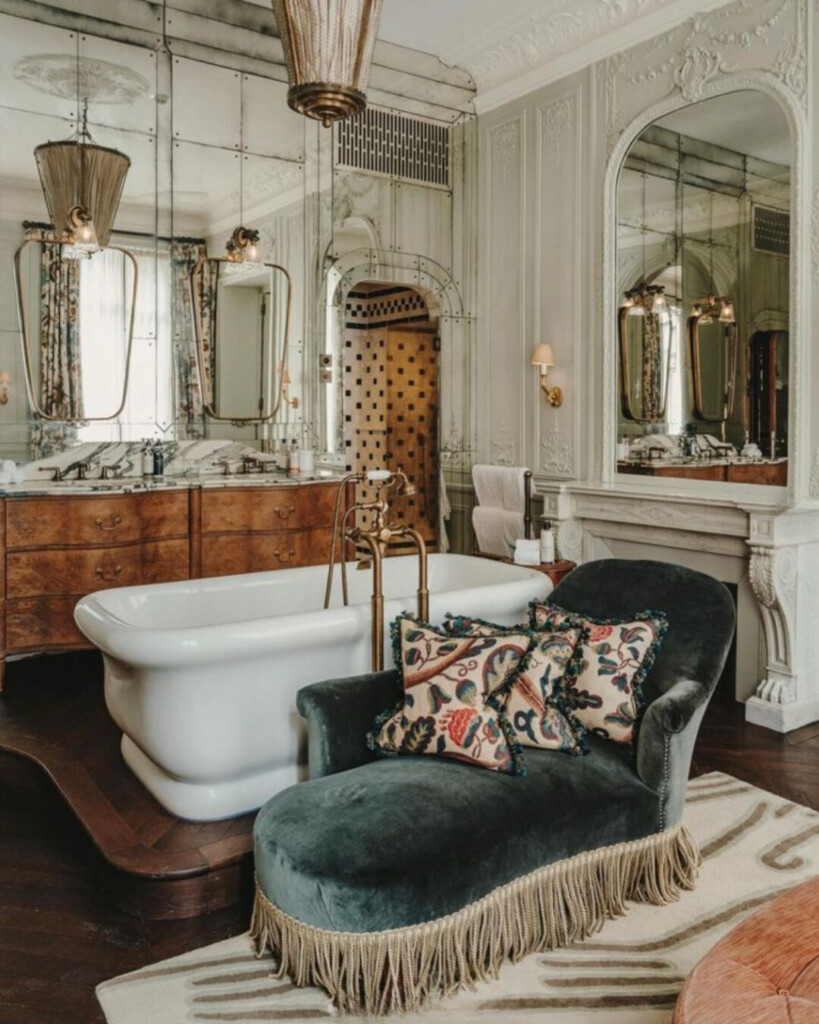 Parisian-style bathroom interior featuring a freestanding soaking tub, antique wood vanity, brass fixtures, mirrored wall panels, and a velvet accent chair with fringe trim.