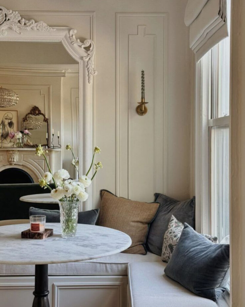 Elegant breakfast nook with banquette seating featuring a marble bistro table, upholstered bench, layered neutral pillows, ornate mirror, and classic wall molding in a Parisian-inspired space.