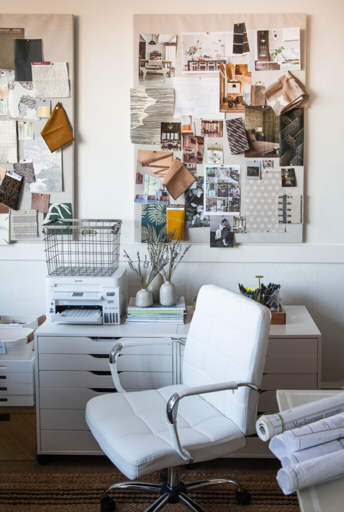 Bright home office with a white desk, modern white swivel chair, printer, stacked design books, and two large inspiration boards filled with fabric swatches, interior photos, and material samples pinned to the wall.