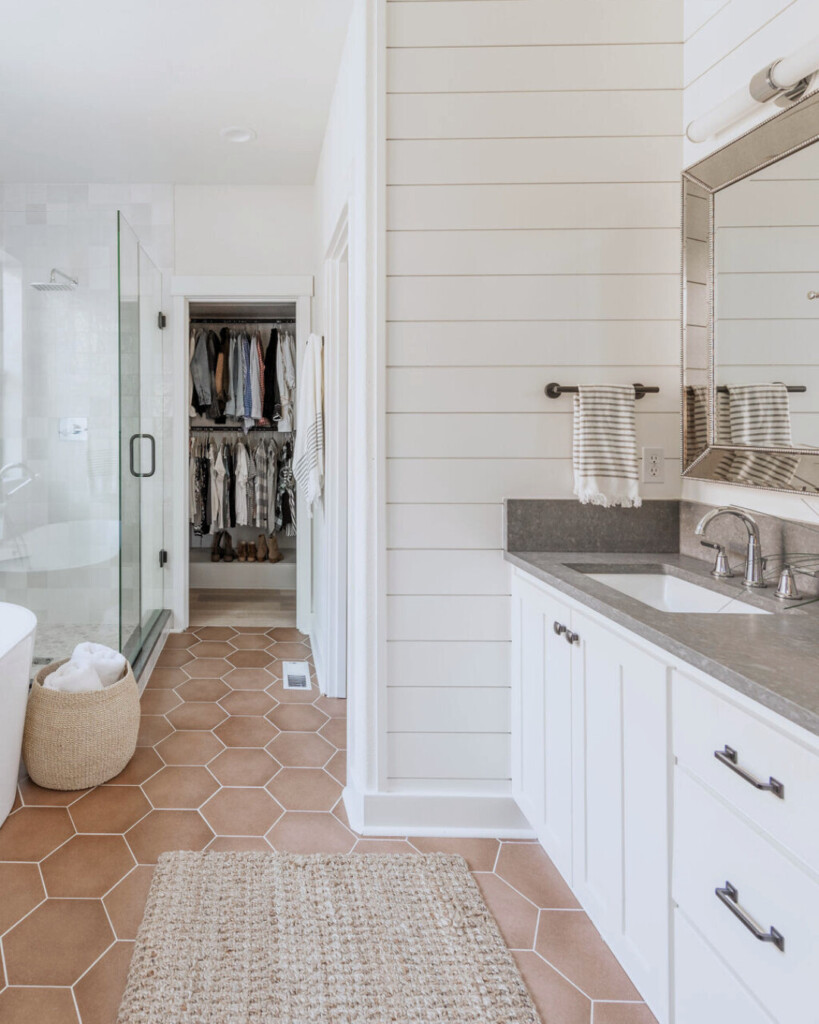 Bright modern bathroom with white shiplap walls, a gray stone vanity, hexagon terracotta tile floors, glass shower, and an open closet visible through the doorway.