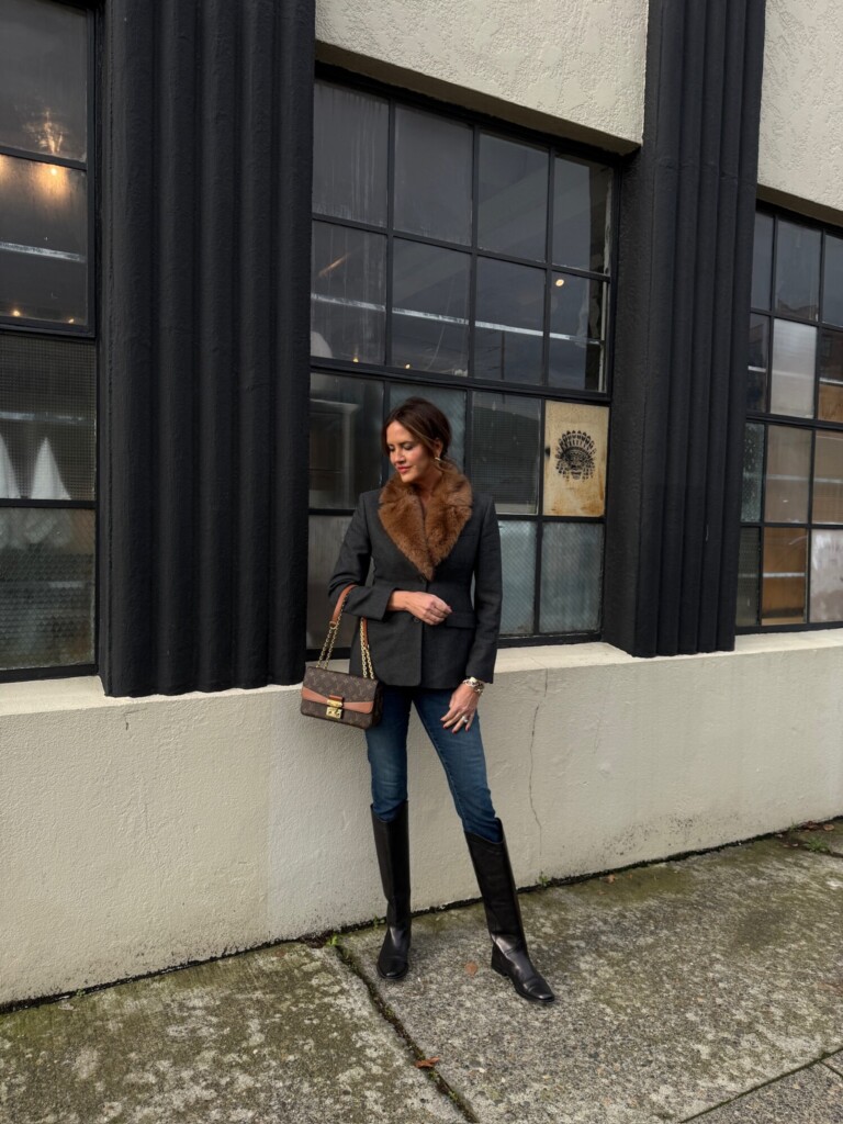 Woman wearing a tailored dark blazer with a brown fur collar, slim blue jeans, and tall black leather boots, standing outside an industrial-style building with black textured columns and large grid windows, styled with a brown monogram shoulder bag.