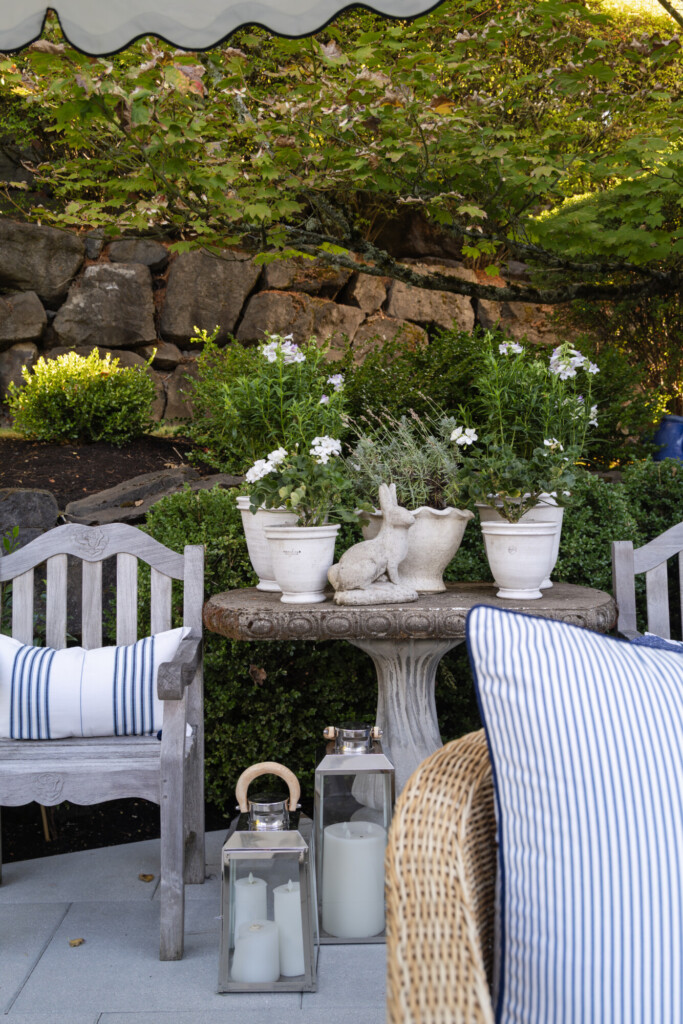 Cozy outdoor patio seating with wooden bench, blue striped pillows, stone pedestal table styled with potted plants, and lantern candles in a lush garden setting