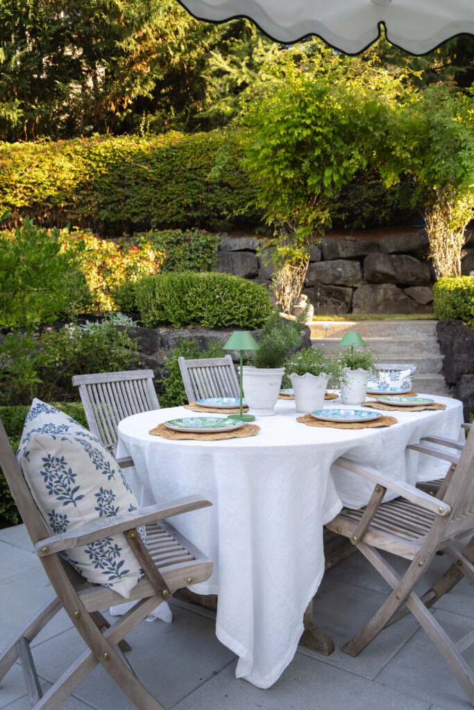 Round outdoor dining table with white linen tablecloth, woven placemats, green cordless lamps, and wooden folding chairs set against lush landscaping and stone retaining walls