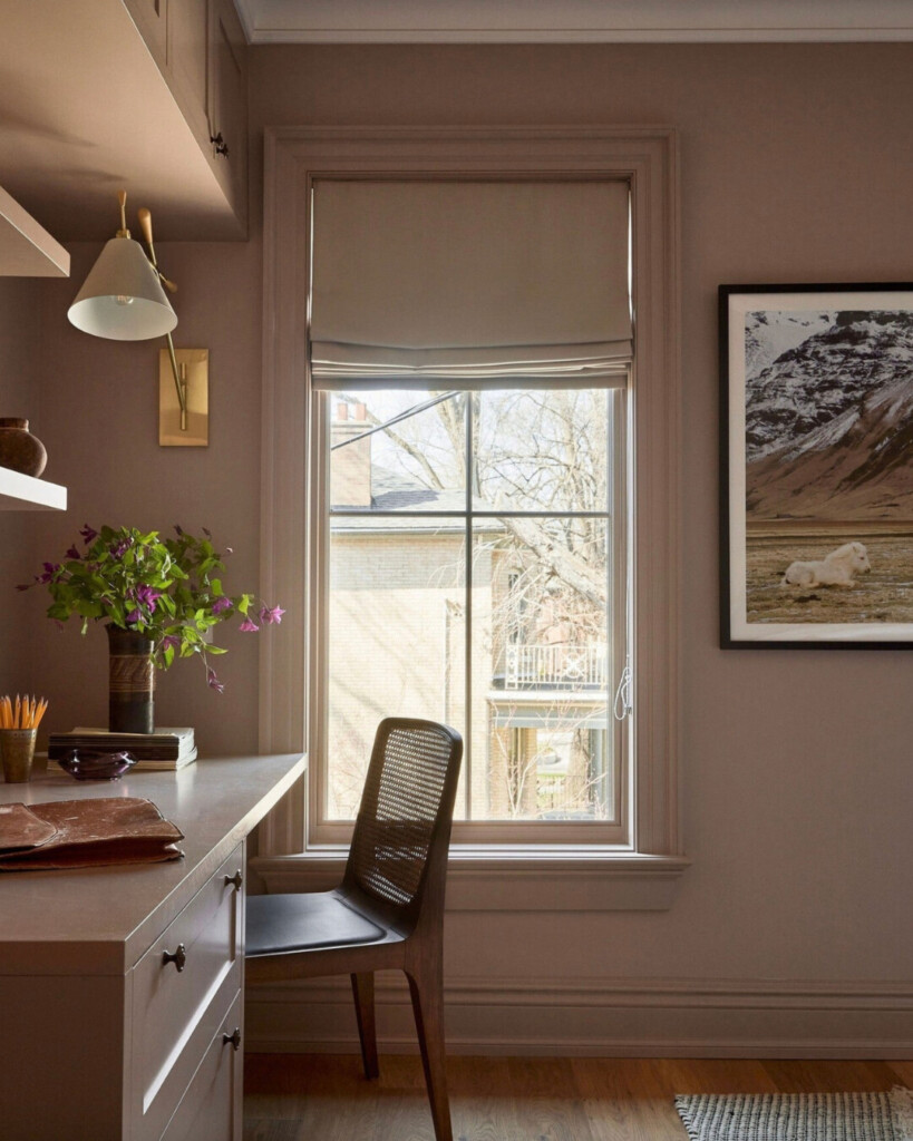 Soft pink home office with built-in desk and shelving, featuring a warm blush wall color, brass wall sconce, woven desk chair, and natural light from a large window.