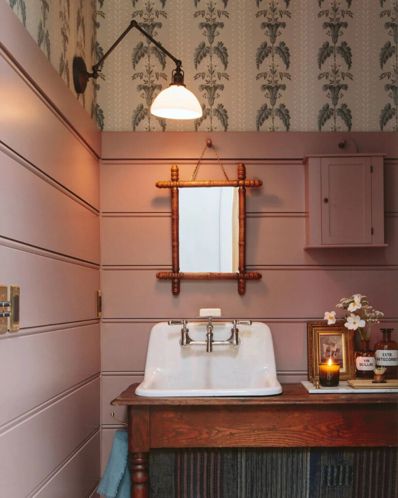 Blush pink bathroom with vertical paneling and patterned wallpaper, featuring a vintage-style sink, brass fixtures, wooden mirror, and warm layered lighting.