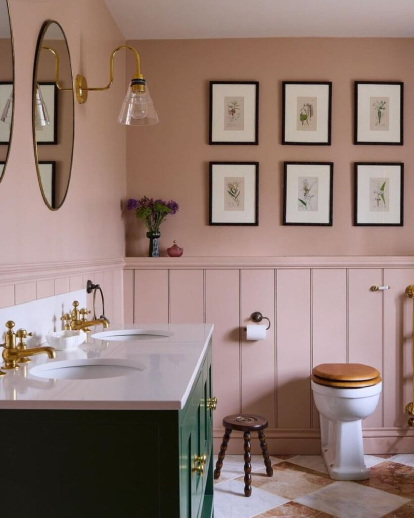 Blush pink bathroom with green vanity and brass fixtures, featuring framed botanical artwork, paneled walls, and classic checker tile flooring.