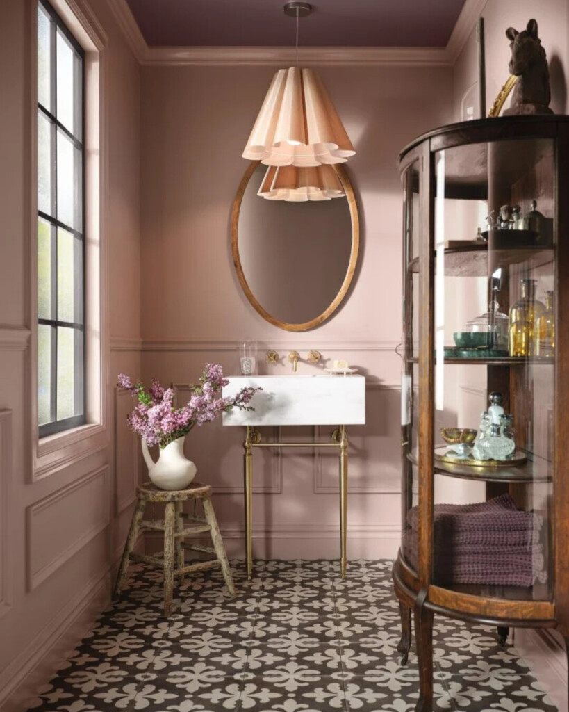 Soft pink powder room with marble sink and patterned tile floor, featuring brass plumbing, a sculptural pendant light, and a glass display cabinet.Blush pink bathroom with green vanity and brass fixtures, featuring framed botanical artwork, paneled walls, and classic checker tile flooring.