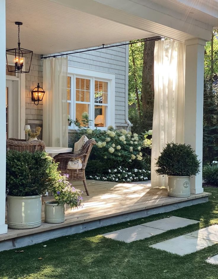 Charming covered porch with wicker seating, sheer outdoor curtains, lantern lighting, and large potted plants overlooking a manicured lawn