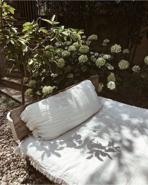 Low outdoor daybed with white cushions set on gravel, surrounded by leafy greenery and blooming hydrangeas in soft dappled sunlight