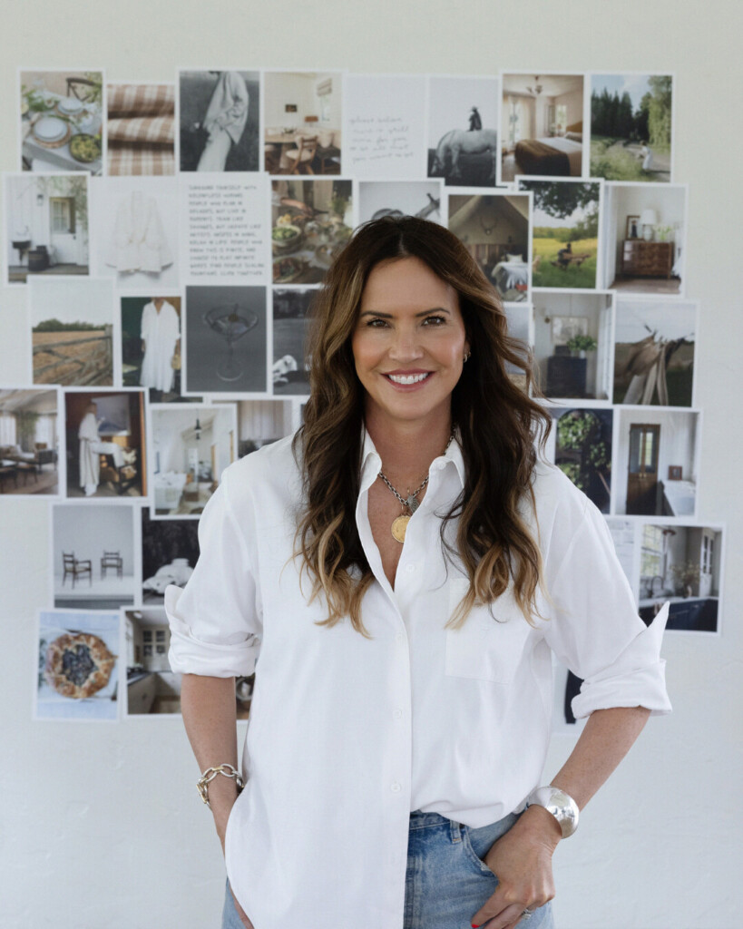 Allison: Woman with long brunette hair wearing a white button-down shirt and jeans, standing in front of a wall collage mood board filled with interior design and lifestyle inspiration images.