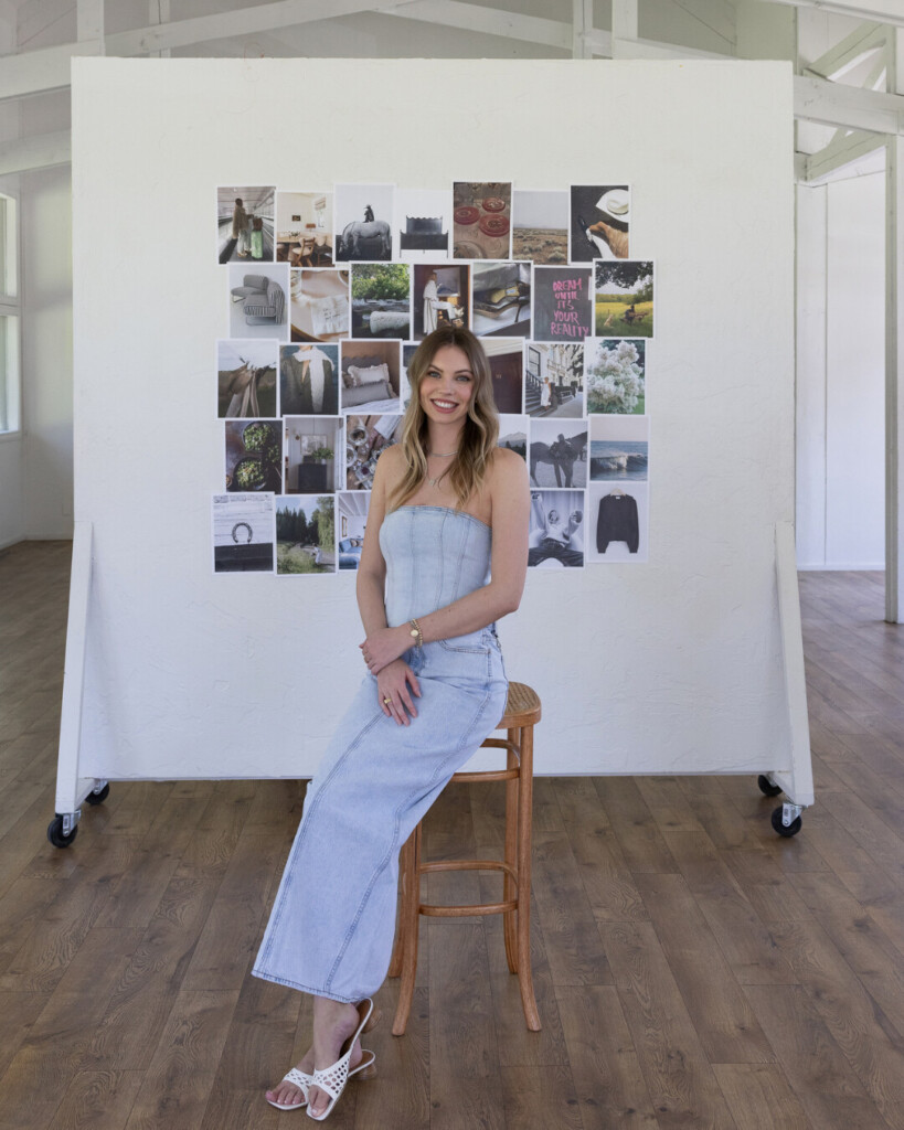 Emma: Woman in a light denim strapless dress sitting on a wooden stool, smiling in front of a white rolling mood board filled with neutral-toned interior and lifestyle inspiration photos.