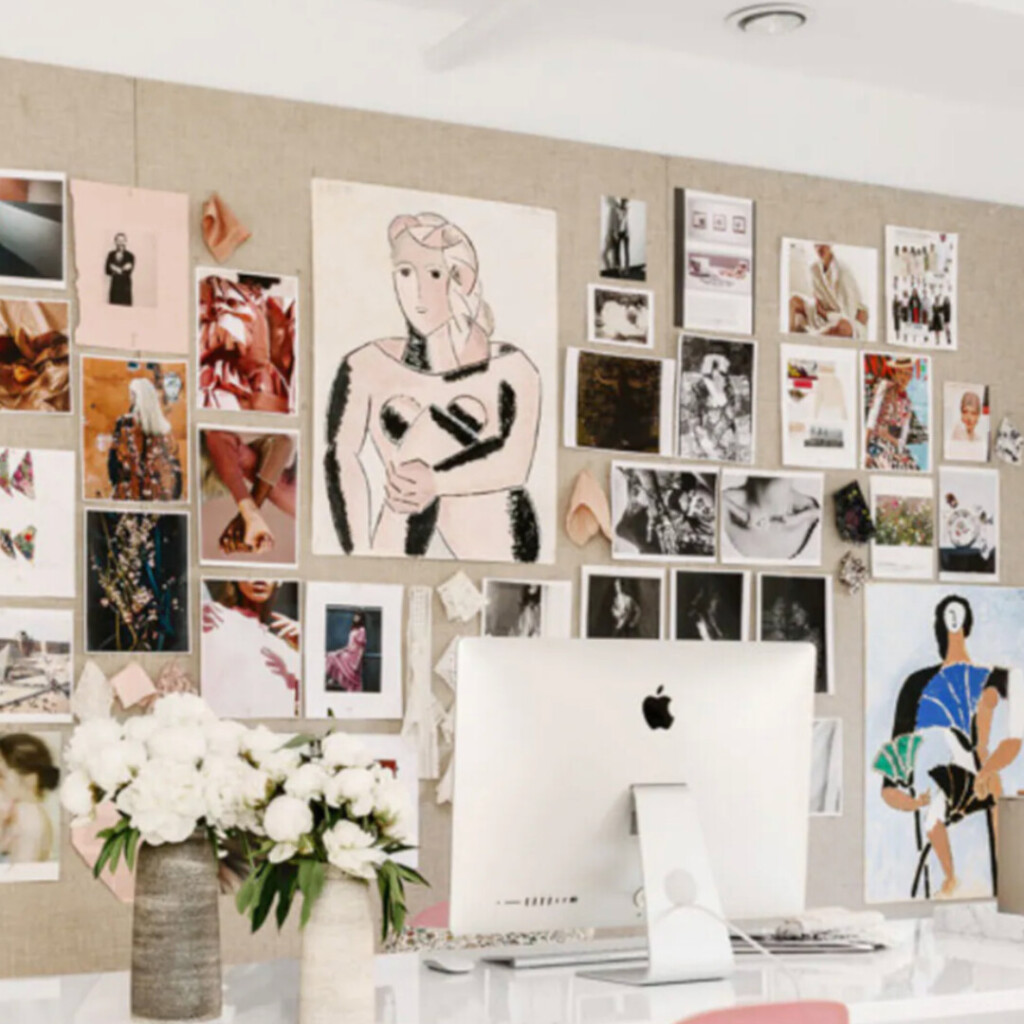 Creative home office with a large inspiration bulletin board filled with art prints, fashion imagery, and sketches above a desk with an iMac, styled with ceramic vases and white flowers for a collected, studio-like feel.