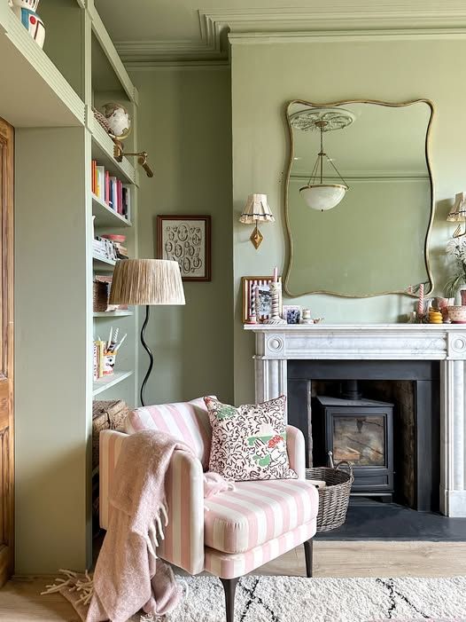 Soft green living room with color-drenched walls and trim, built-in shelving, a fireplace, striped accent chair, patterned Roman shade, and layered textures creating a calm, collected, lived-in space.