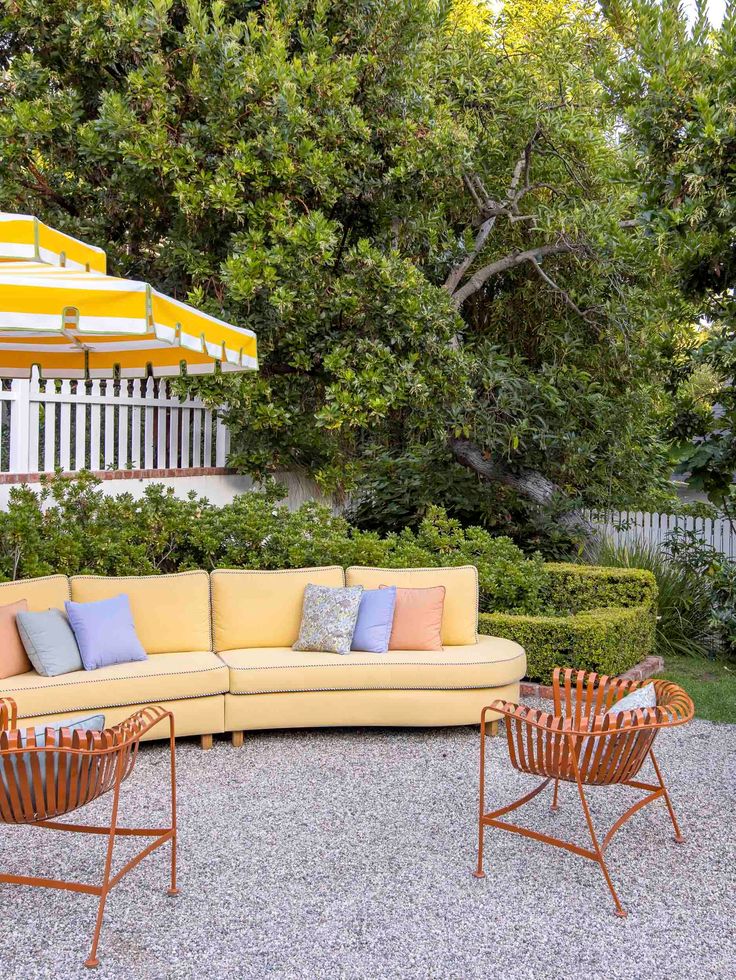 Sunny outdoor seating area with a yellow sofa, rust metal lounge chairs, striped umbrella, and manicured hedges set on a gravel patio surrounded by lush greenery
