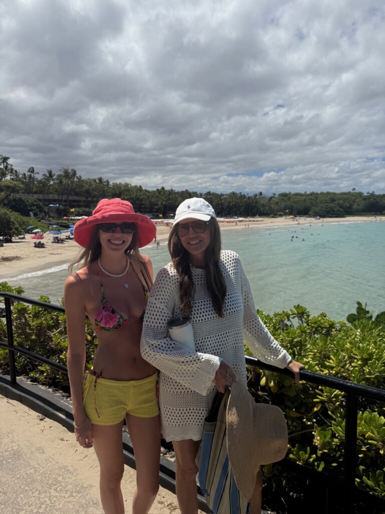 Mother and daughter smiling by the ocean on a sunny beach day, wearing hats and casual beachwear, with turquoise water, sandy shoreline, and palm trees in the background.
