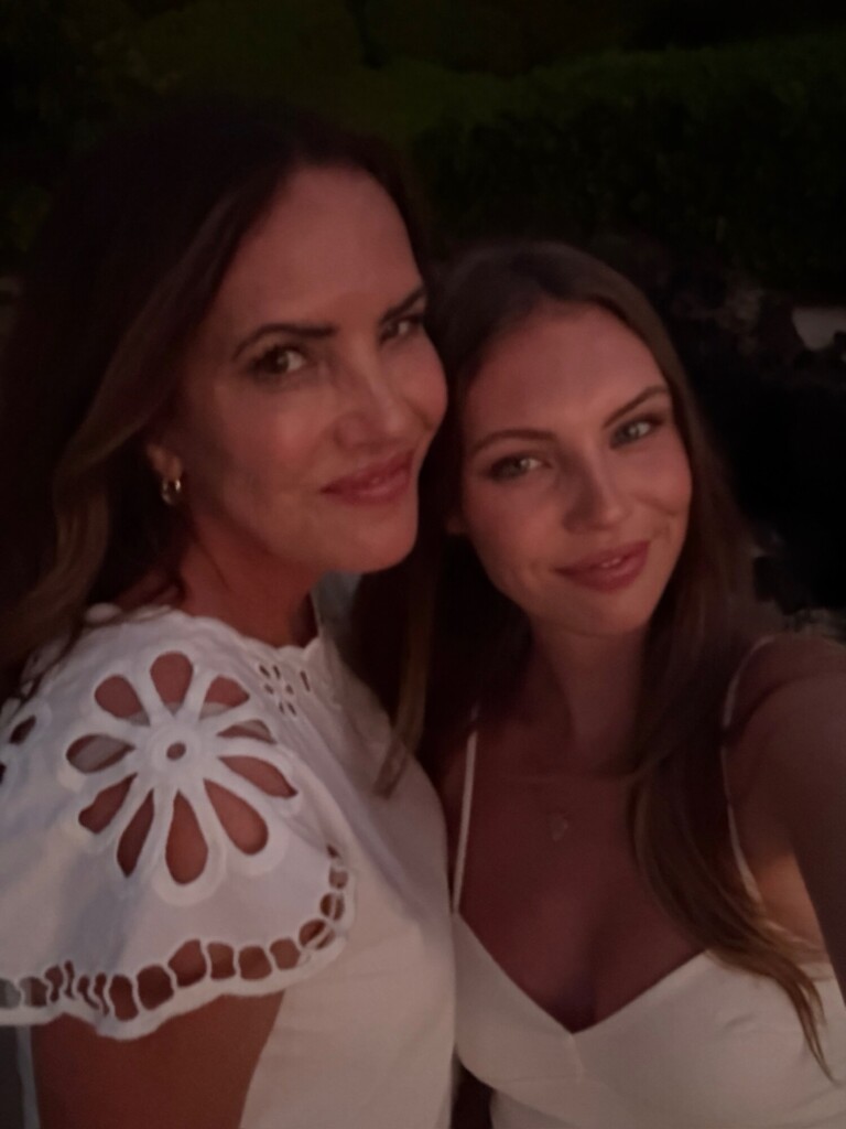 Mother and daughter taking a sunset selfie outdoors, both wearing light-colored dresses and smiling, with warm evening light and greenery in the background.