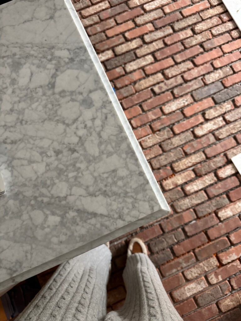 Top-down view of a marble countertop edge meeting warm, over-grouted brick veneer kitchen flooring, showing contrast between smooth stone and textured brick.