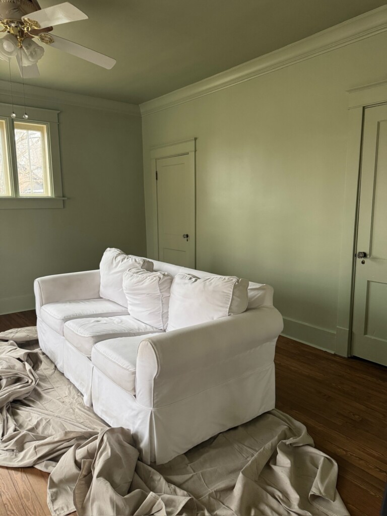 A soft green room painted in a muted Vert de Terre–style hue, featuring a white slipcovered sofa centered on warm wood floors. Classic trim details, a simple ceiling fan, and painted doors blend seamlessly into the walls, creating a calm, cohesive backdrop with an understated, timeless feel.