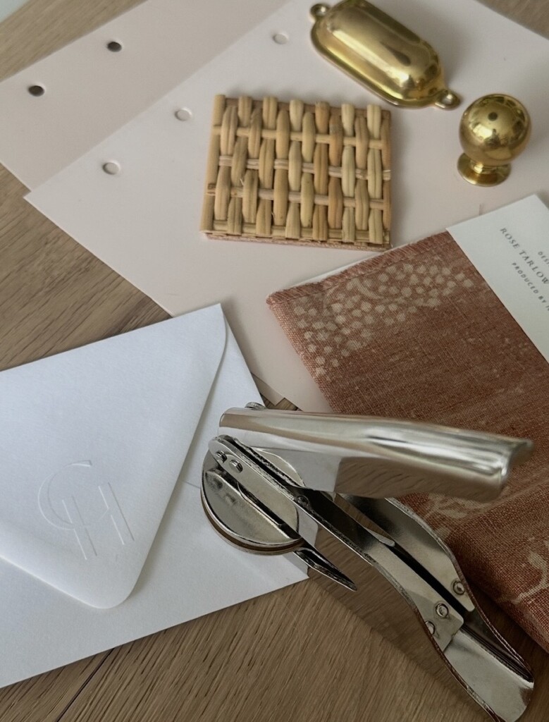 A flat-lay of interior design materials arranged on a light wood surface, including fabric swatches in warm blush and neutral tones, a woven rattan sample, brass hardware pieces, paint sample cards, and a metal hole punch resting on a white envelope. The composition highlights texture, material contrast, and a curated, behind-the-scenes design process.