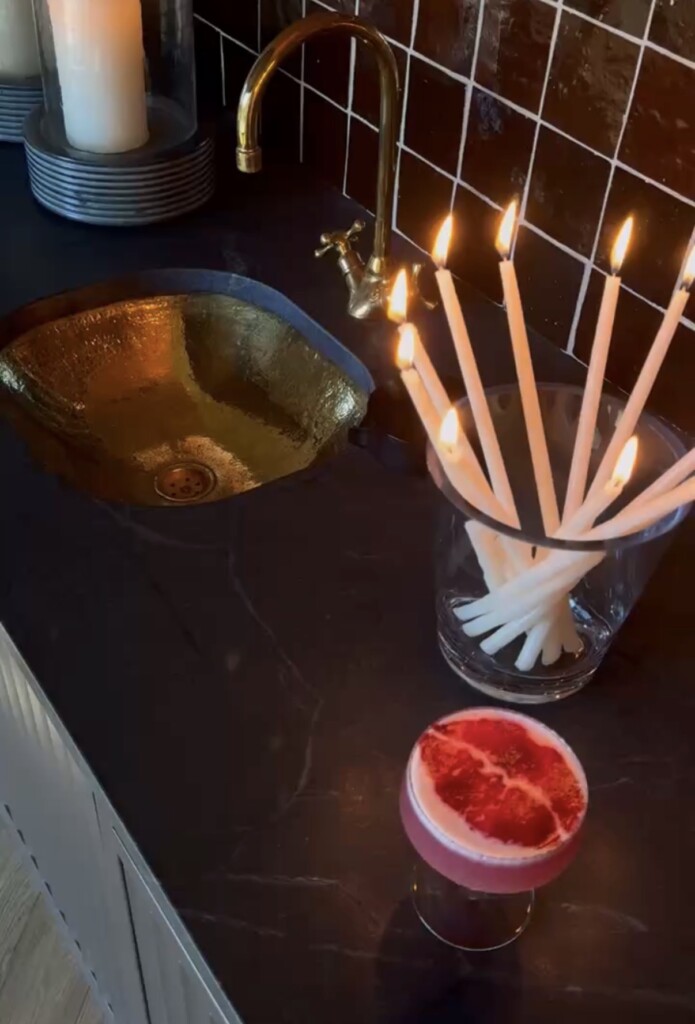 A moody bar setup with a hammered brass sink and vintage-style brass faucet set into a dark countertop, a grid-patterned brown tile backsplash, and a glass filled with lit white taper candles. In the foreground sits a raspberry-colored cocktail topped with foamy pink froth, creating a cozy, intimate, candlelit atmosphere perfect for entertaining.