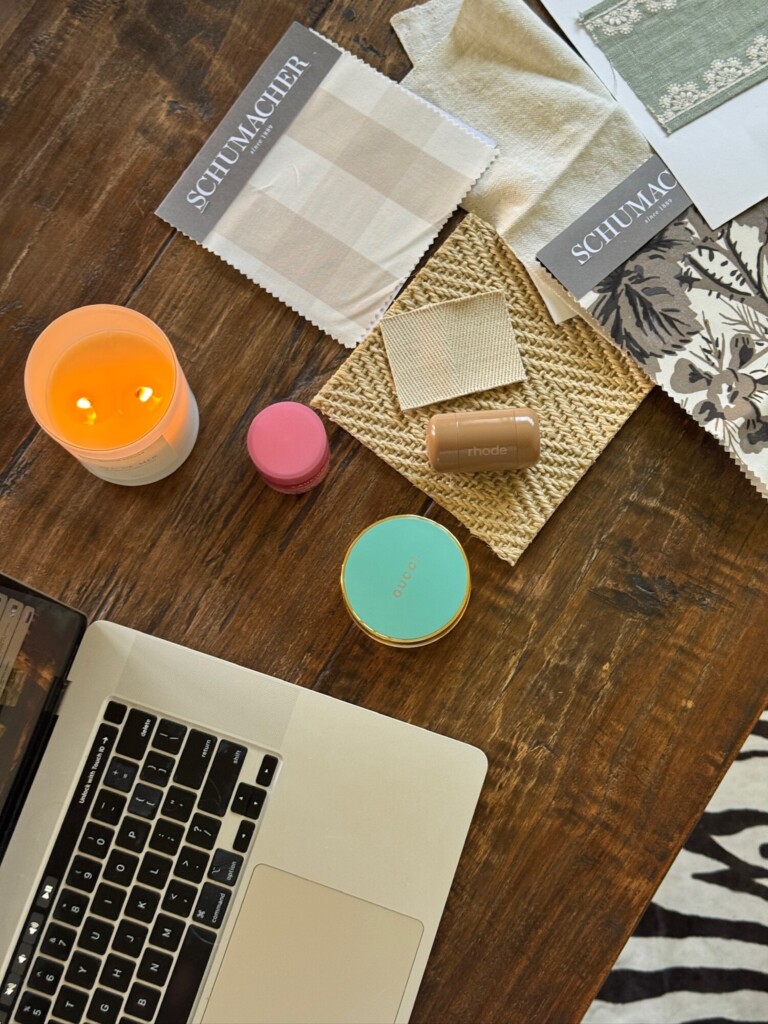 Flat lay of a wooden desk featuring interior design fabric swatches, woven material samples, a lit candle, beauty products, and a laptop, styled in warm neutral tones with natural textures.