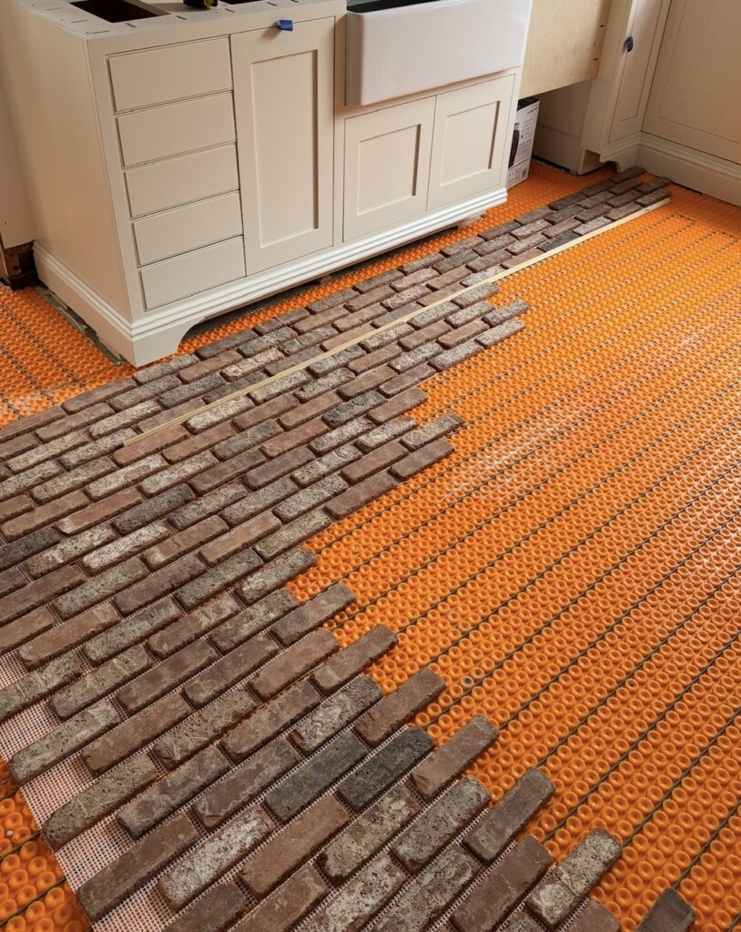 Brick veneer tile installation in progress over radiant heated floor mats, showing staggered brick layout beneath custom kitchen cabinetry.