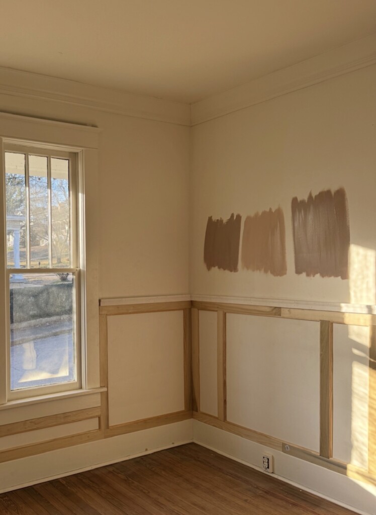 Corner of a sunlit room with newly installed natural wood wainscoting and three warm brown paint swatches brushed across a cream wall above the trim. Late afternoon light casts long shadows across the wall and hardwood floors, highlighting the wood detailing.