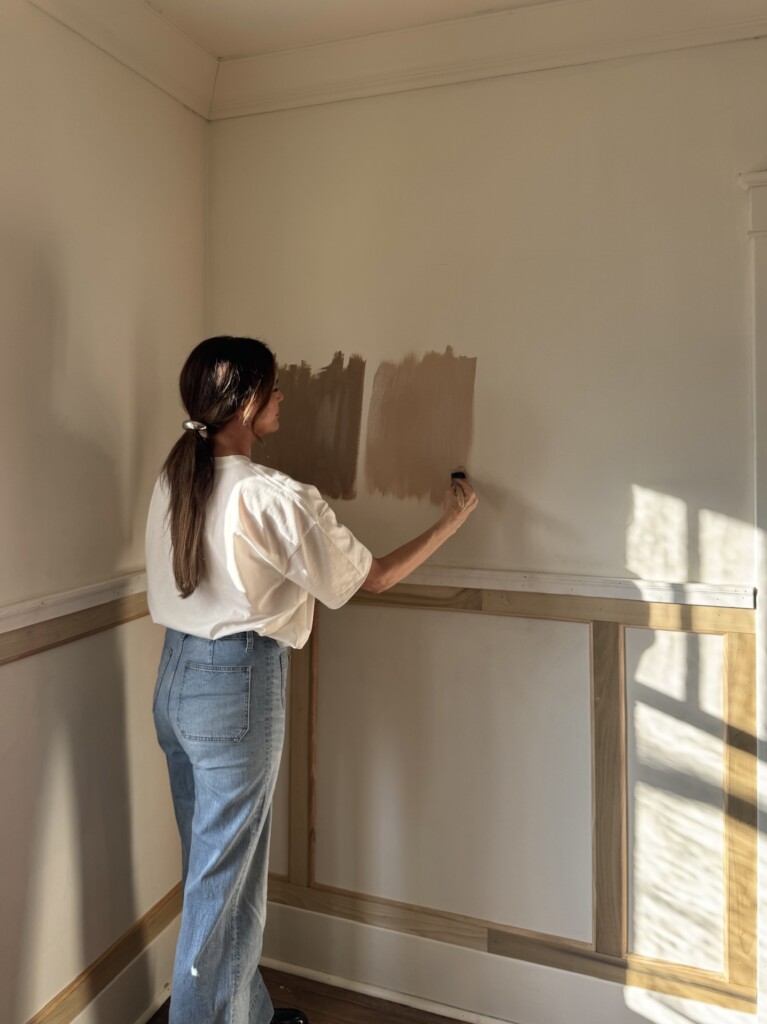 Woman testing warm brown paint samples on a cream wall above natural wood wainscoting in a bright room. She stands in profile wearing a white tee and light denim, brushing paint onto the wall as golden sunlight streams through a nearby window and casts soft shadows.