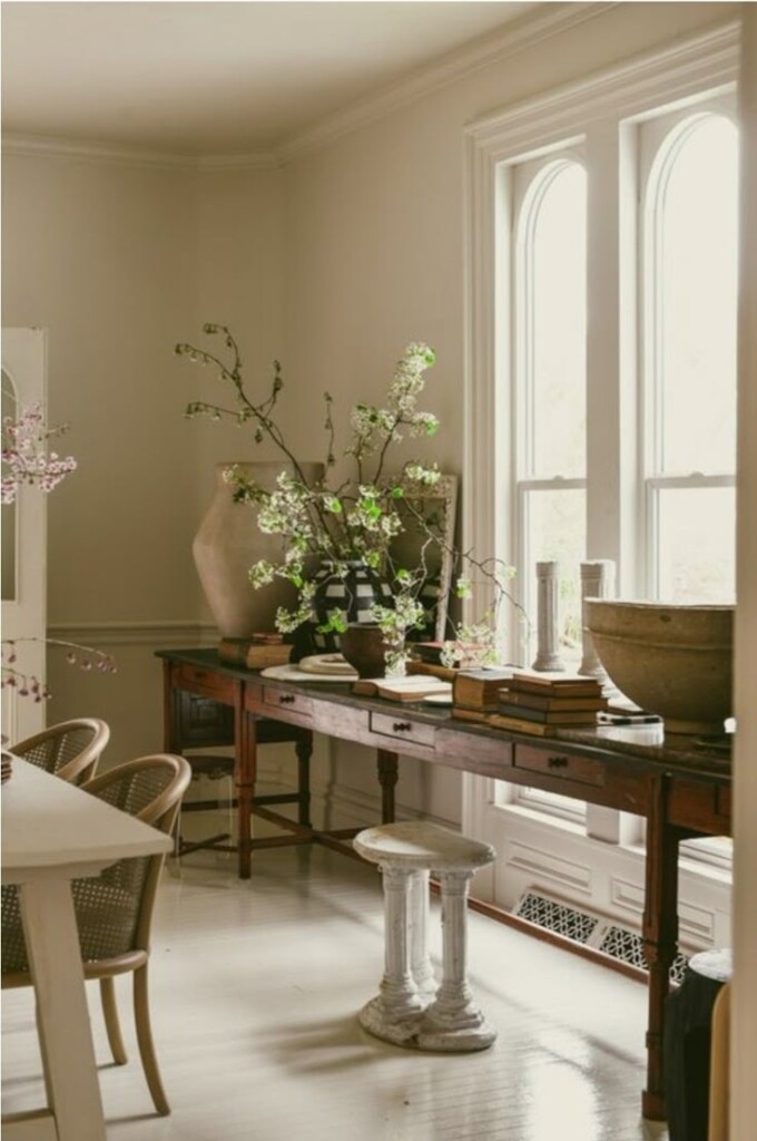 Soft, neutral-toned dining room with tall arched windows, a vintage wooden console table styled with stacked books, pottery, and flowering branches, creating an airy, collected, old-world atmosphere.