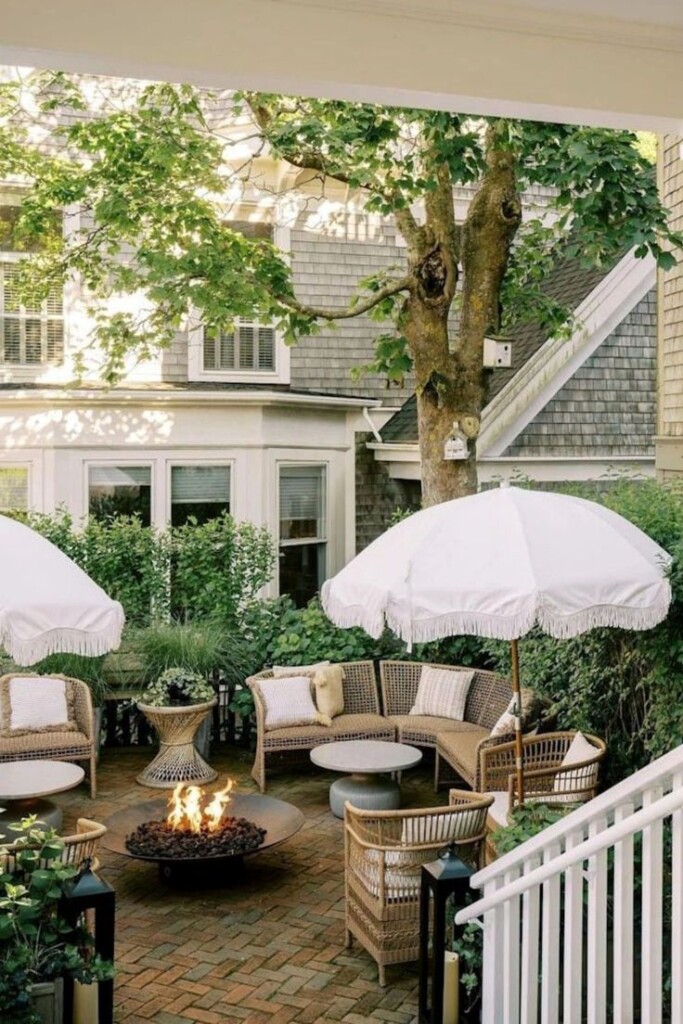 Charming brick patio with wicker seating, white fringe umbrellas, round fire pit, and lush greenery creating a cozy courtyard outdoor living space