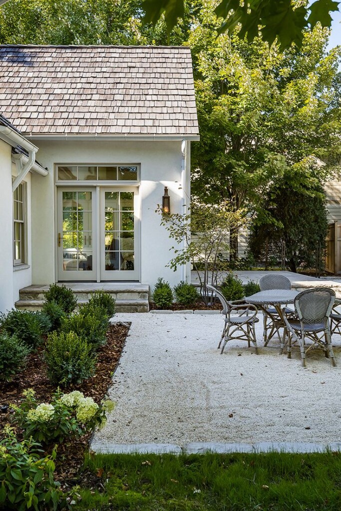 Cozy side yard patio with pea gravel ground cover, French doors, bistro dining set, and neatly trimmed boxwood landscaping beside a white cottage-style home