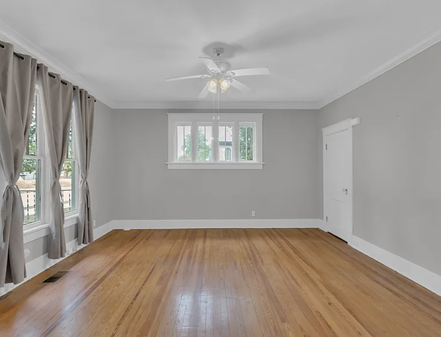 Empty room with light gray walls, white trim and crown molding, warm hardwood floors, a white ceiling fan with light, simple gray curtains on tall windows, and a small central window creating a clean, neutral space ready for transformation.