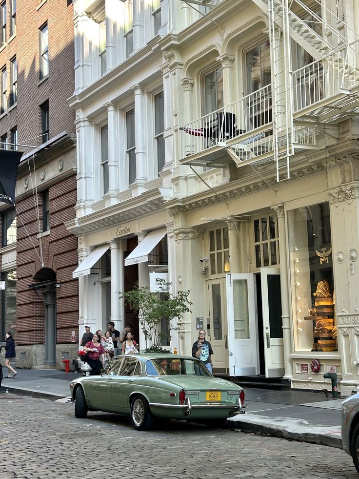 SoHo street scene in New York City featuring classic cast-iron architecture, white fire escapes, boutique storefronts, cobblestone street, and a vintage green car parked along the curb.