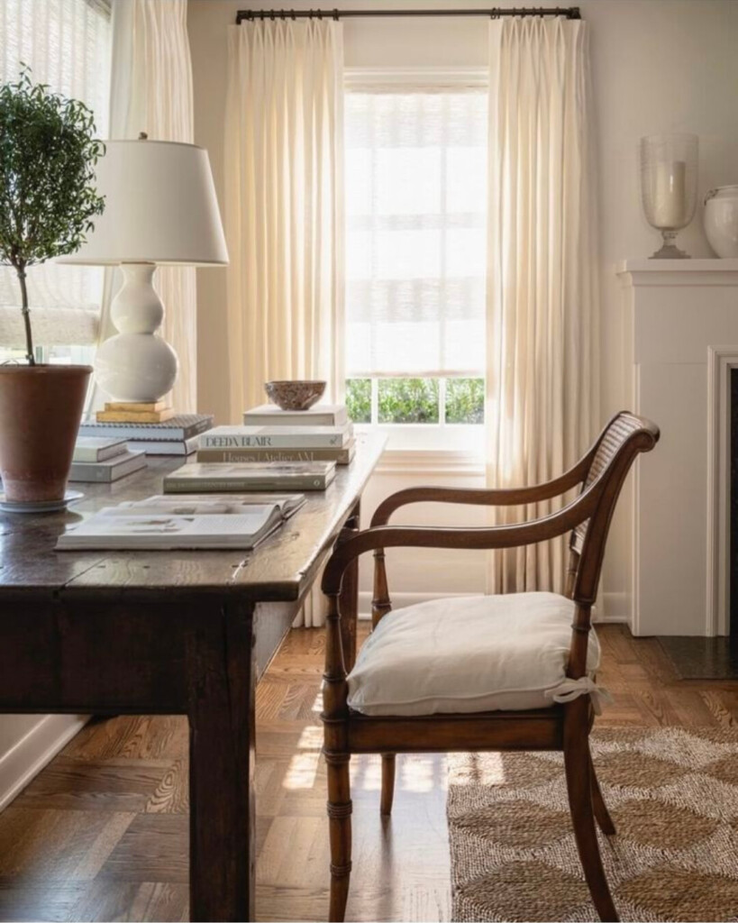 Warm, light-filled home office with sheer linen curtains, a rustic wooden desk, a classic wood armchair with a cushion, stacked books, and a table lamp, styled with natural textures and an old-world, collected feel.