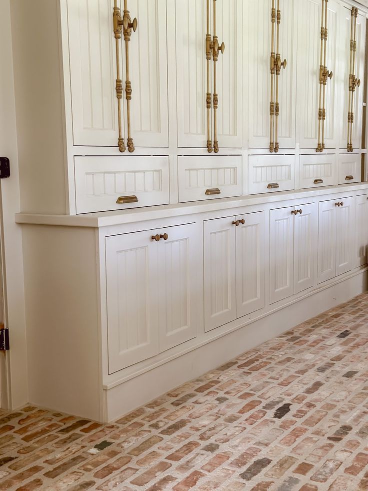 White built-in kitchen cabinetry with beadboard paneling and antique brass hardware, paired with warm brick veneer flooring laid in a staggered pattern for a timeless, old-world cottage feel.