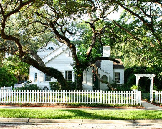Charming white cottage-style home with picket fence, mature oak trees, and arched garden arbor along a tree-lined street