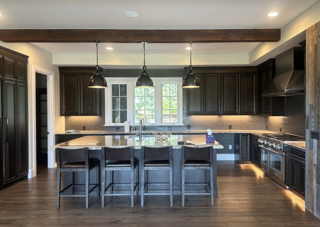 Wide view of a dark mahogany kitchen in a timber home with large island, granite countertops, black pendant lights, and exposed wood beam ceiling in Bend, Oregon.