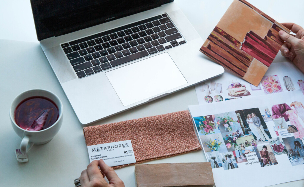 Workspace flat lay with a laptop, fabric swatch, inspiration photos, and tea on a desk used for interior design planning and material selection.