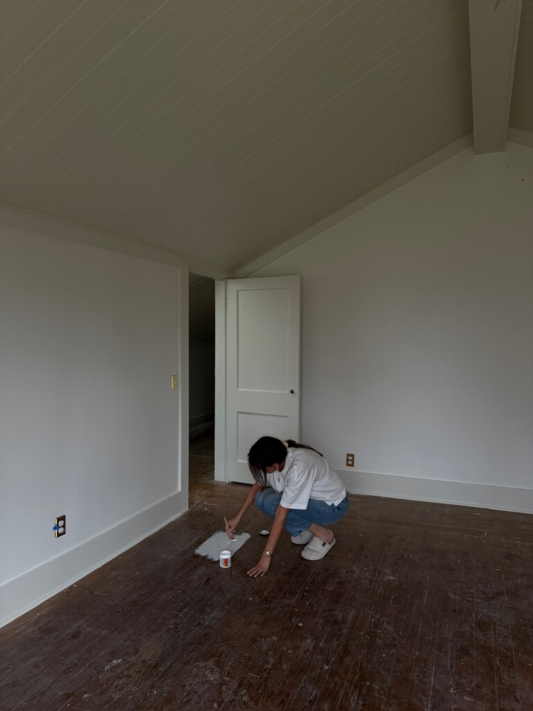 Person kneeling on worn wood floors testing paint samples; small swatches of creamy white tones applied as natural light fills the room.