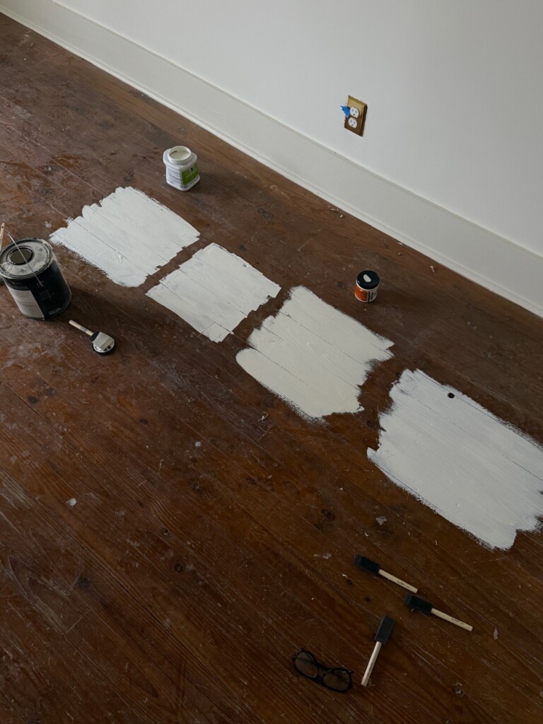 Close-up of wood floor with multiple white paint samples, paint cans, brushes, and tools laid out, showing the process of selecting the right floor color.