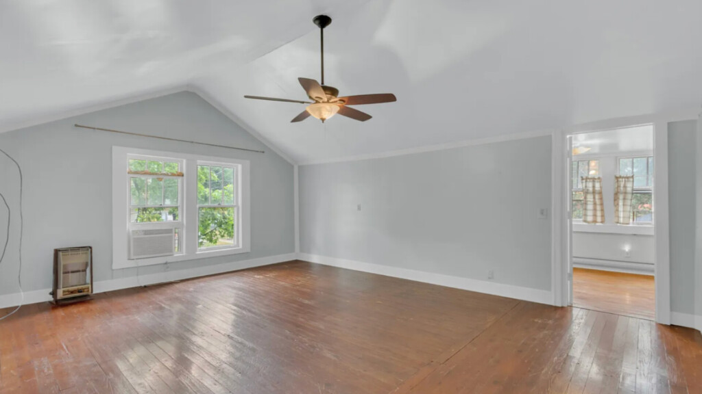 Empty upstairs bedroom with vaulted ceiling, light gray walls, white trim, and worn wood floors; double windows with greenery view and window AC unit; ceiling fan overhead; doorway leading to adjacent room with patterned curtains.