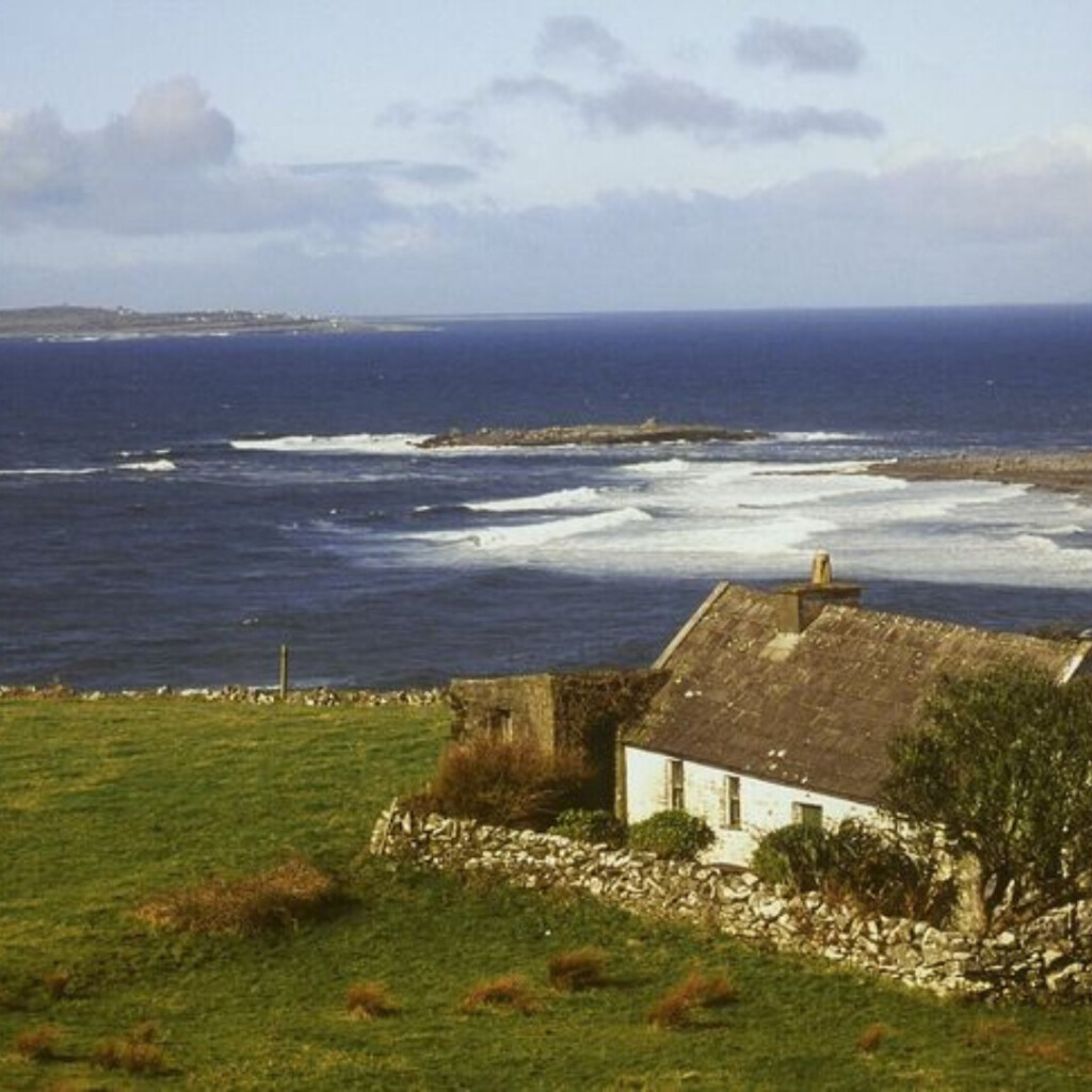Coastal Irish countryside with a small stone cottage overlooking the ocean, rolling green fields, rugged shoreline, and waves crashing along the rocky coast under a cloudy sky.