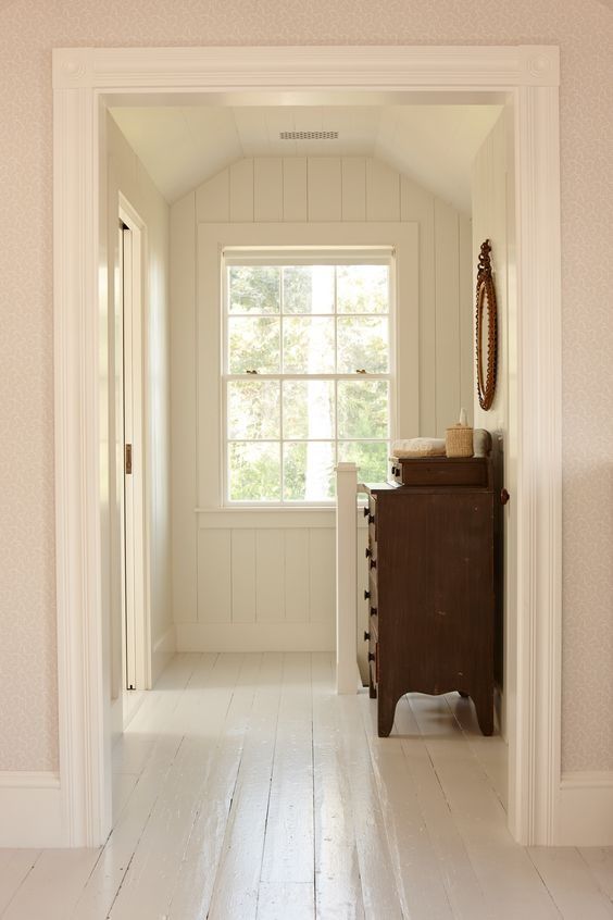 Narrow hallway with painted white wood floors, paneled walls, and a large window at the end; antique wooden dresser styled simply beneath soft natural light.