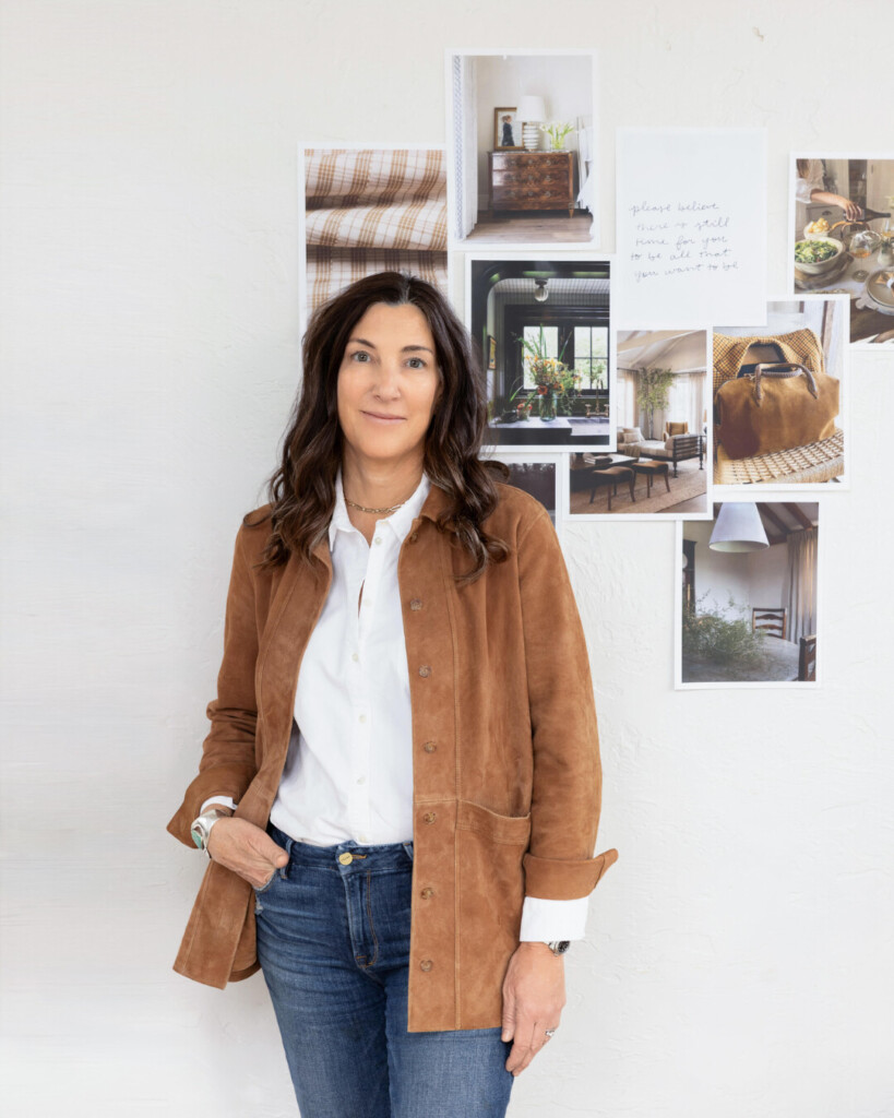 Sarah: Woman with long brunette hair wearing a white button-down shirt and jeans, standing in front of a wall collage mood board filled with interior design and lifestyle inspiration images.
