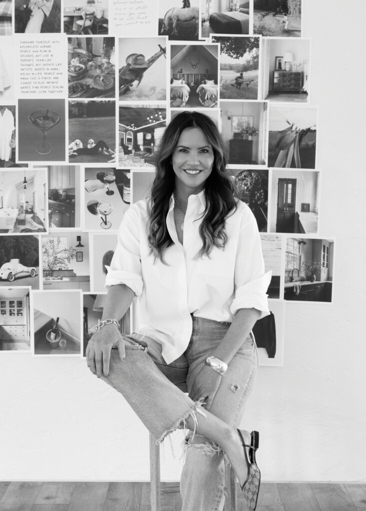 Interior designer seated on a stool in front of a large inspiration mood board filled with black-and-white interior images, lifestyle photography, and design references.