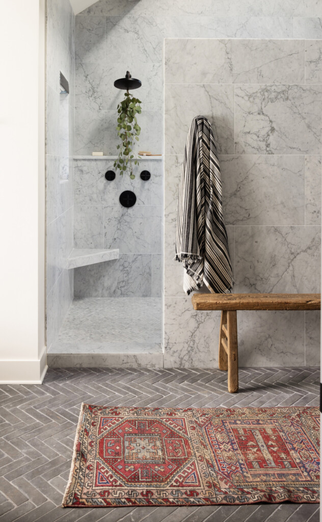 Marble walk-in shower with black fixtures, built-in shelf, trailing plant, and a rustic wooden bench on gray herringbone tile flooring.