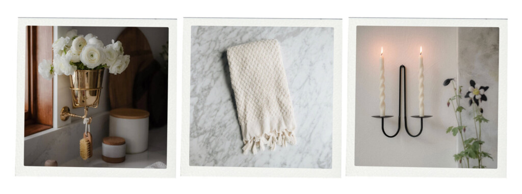 A styled vignette featuring a brass wall-mounted vase with white flowers, a textured cream hand towel on marble, and a black wall sconce with two lit taper candles.