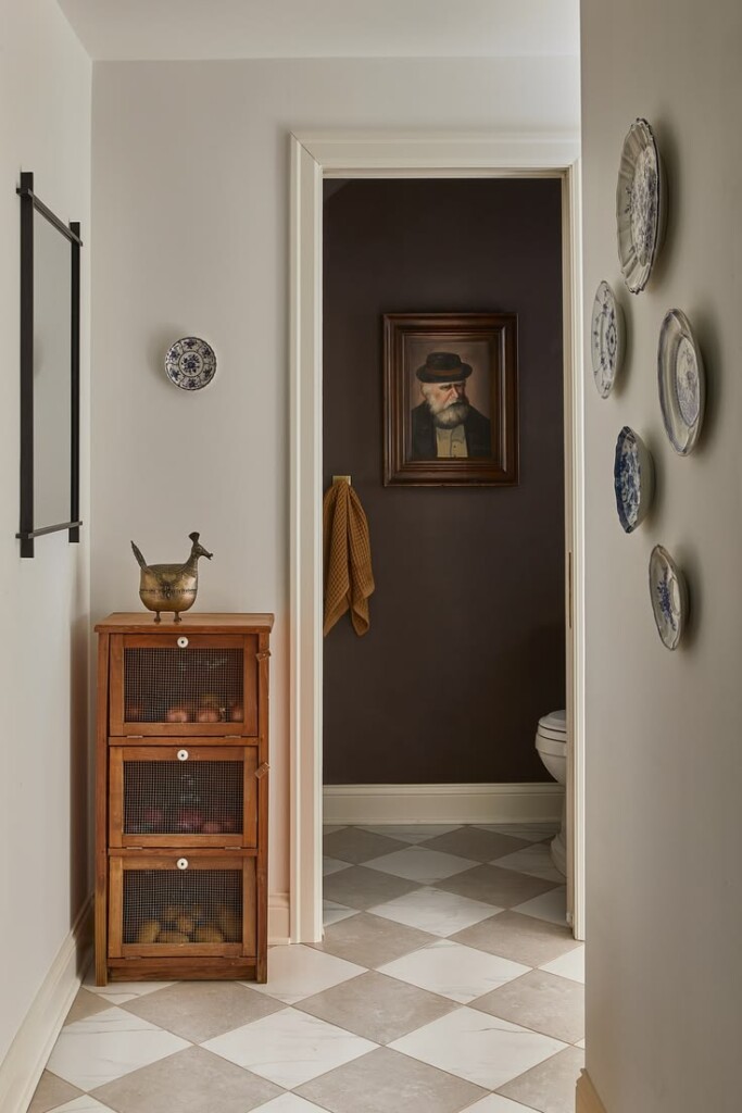 Hallway with cream and tan checkerboard tile floor, small wooden cabinet with mesh drawers, wall-mounted plates, and a framed portrait in a darker adjoining room.