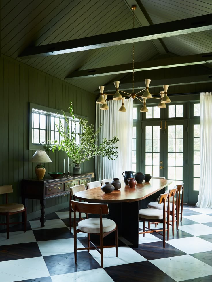 Dining room with deep green paneled walls, exposed ceiling beams, a modern chandelier, and a large black-and-white checkered floor beneath a wooden table.